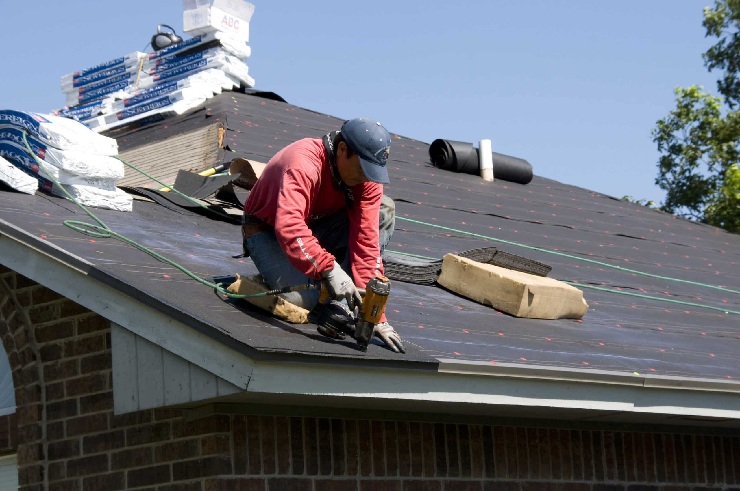 Roofer nailing shingles over black felt underlayment, bundles stacked near the ridge of a brick house