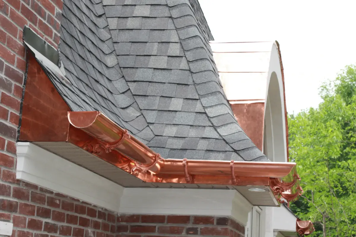 Ornate copper half-round gutter with scrolled cast brackets along a gray shingle eave above a red brick wall