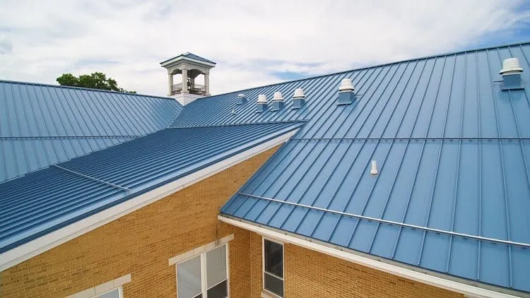 Blue standing-seam metal roof with a cupola and roof vents over a tan brick building