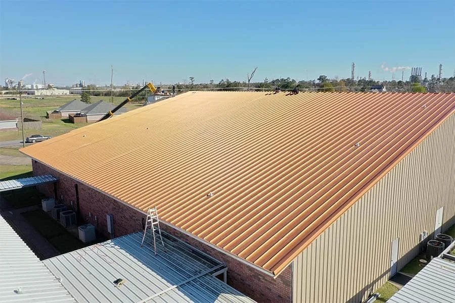 Crew installing a tan corrugated metal roof on a brick commercial building, with a boom lift at the eave
