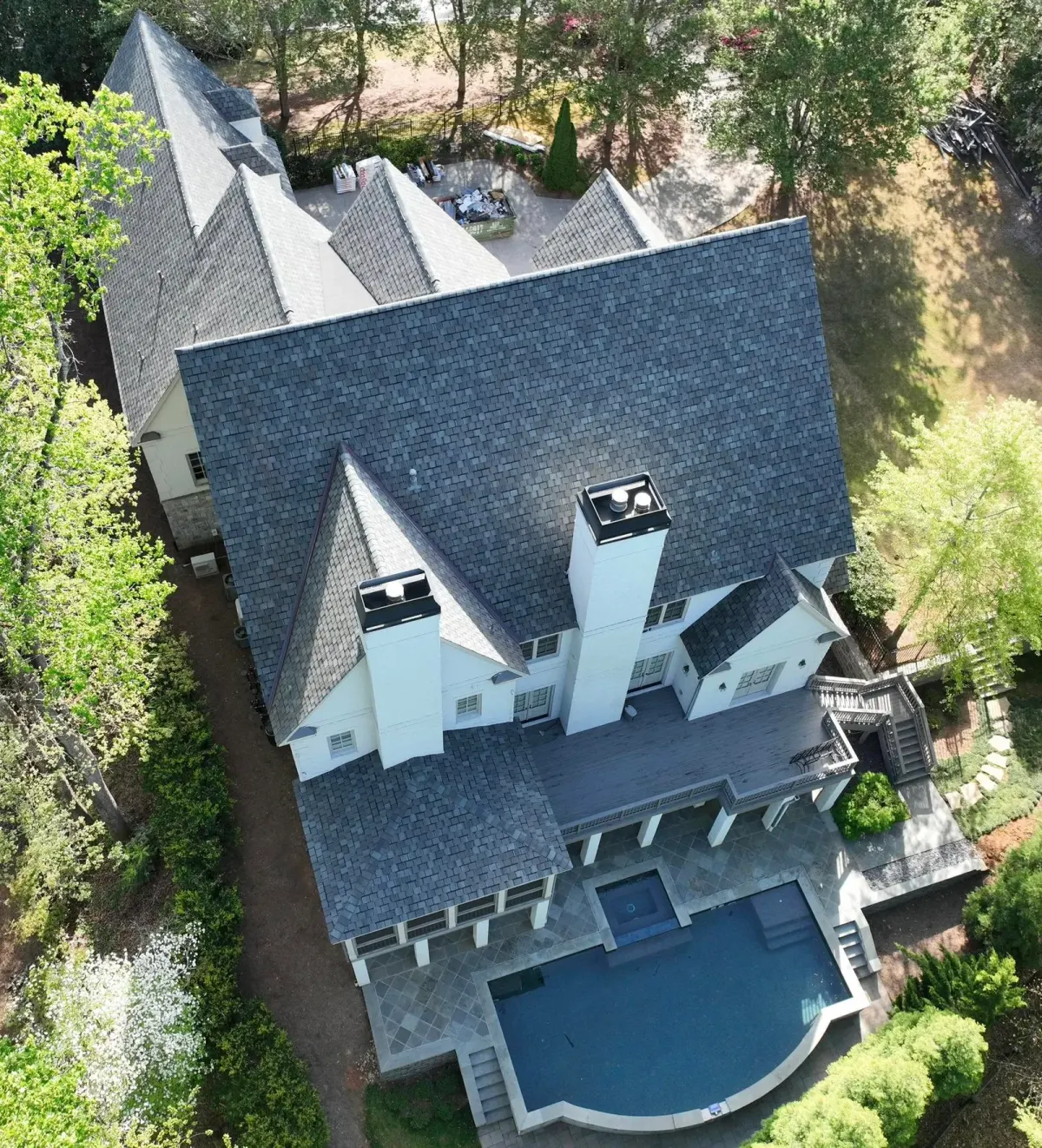 Overhead view of a large gabled home reroofed in gray shingles, tear-off dumpster and new bundles staged out back
