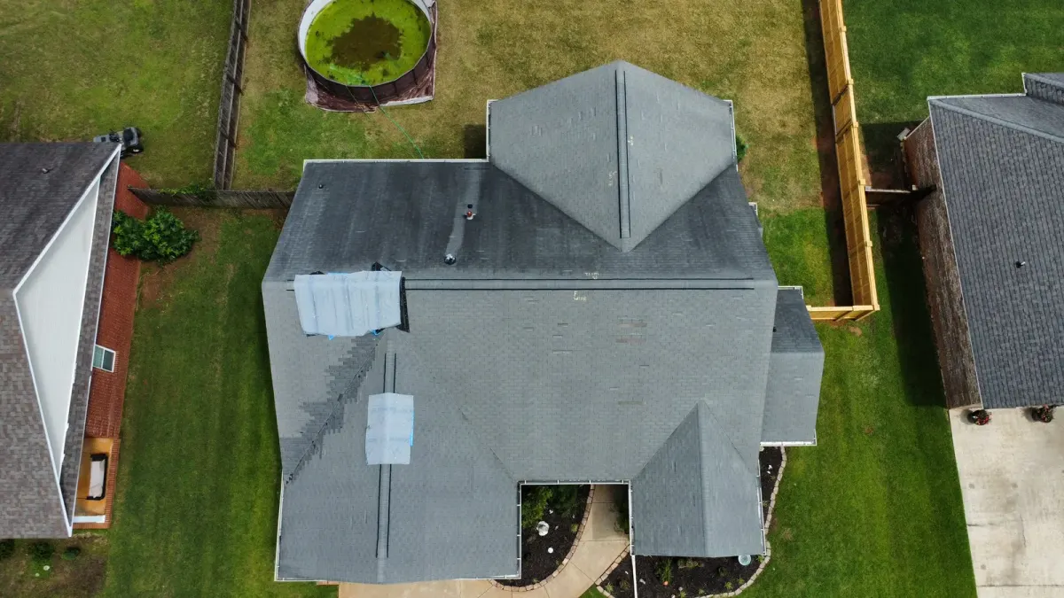 Overhead drone view of a suburban home's gray shingle roof, with white underlayment sheets patched over two torn sections